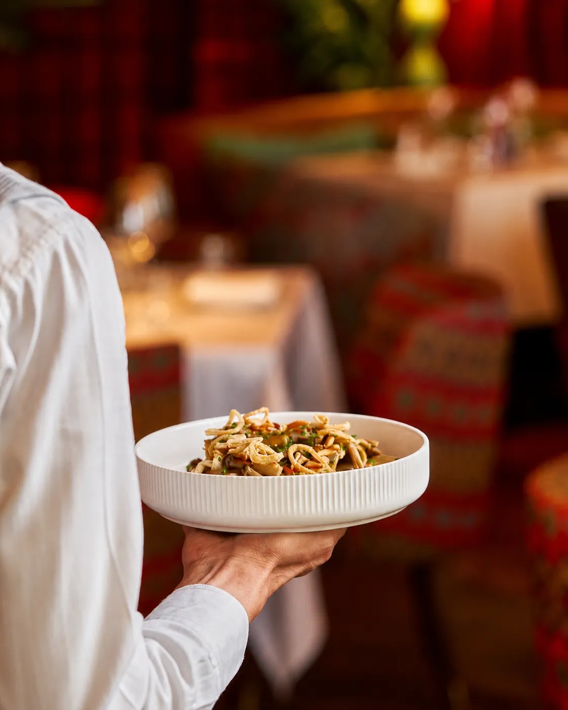A server holding a bowl of pasta in one hand as it is carried through a warmly lit dining room with blurred tables and place settings, conveying an inviting, relaxed atmosphere